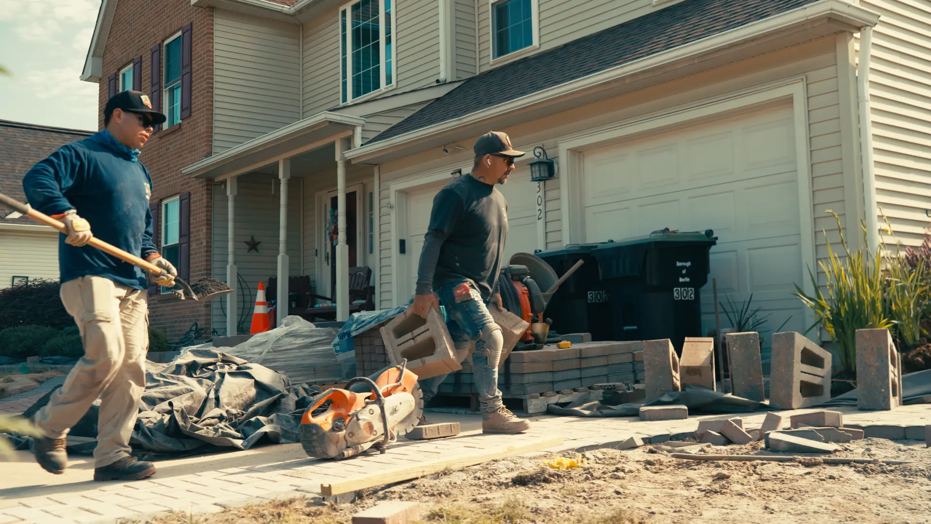 Two crew members hauling wall block and shoveling base material on a front yard paver job in Berlin NJ